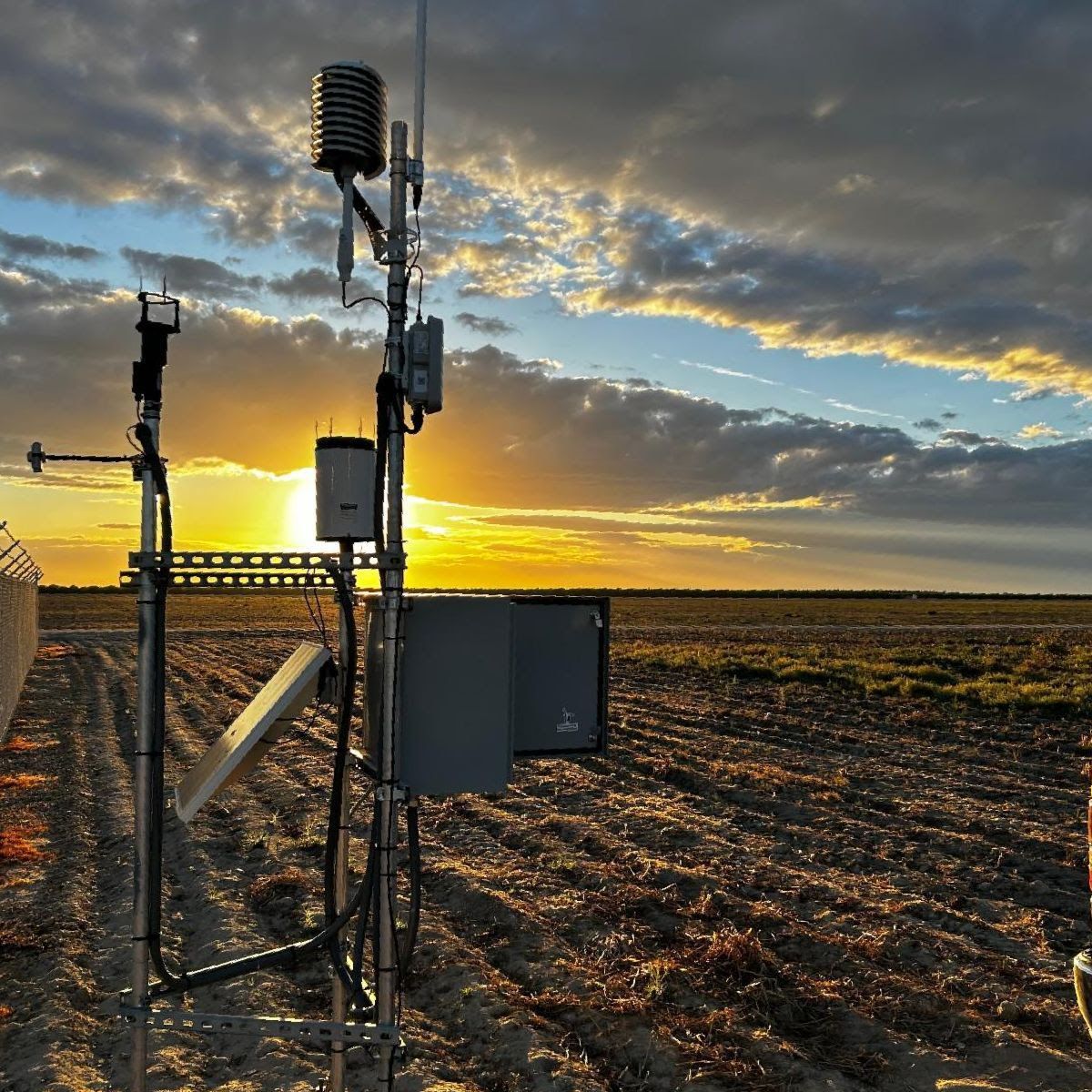 Weather station mast with sensors in a ploughed field at sunset